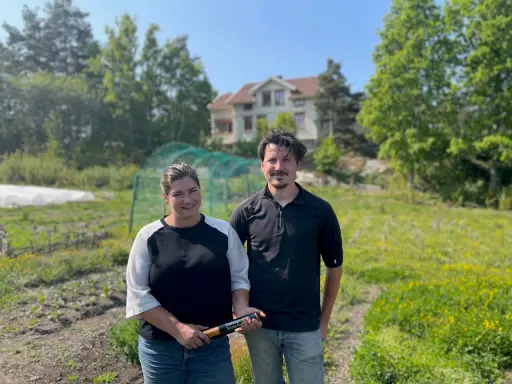 Två personer står på ett fält med ett hus i bakgrunden. De arbetar med lbk lantbruk och visar upp en produkt.