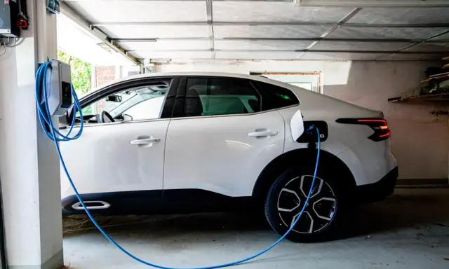 Vit elbil laddas i garage. En blå laddningskabel går från väggen till bilen. Tänk på brandrisk vid laddning av elbil i garage.