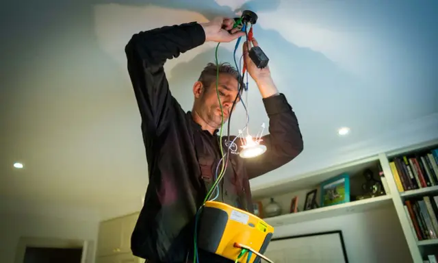 En auktoriserad elektriker kopplar in en ny taklampa. Han har en gul mätutrustning runt halsen.