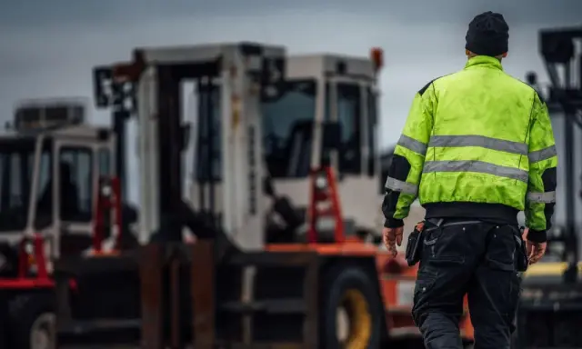 Arbetare i varseljacka inspekterar en truckladdning med flera gaffeltruckar i bakgrunden.