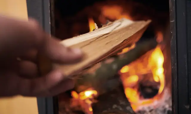 En hand lägger ved i en kamin där elden flammande ryker in från.