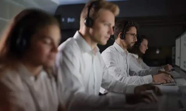 Personal som arbetar på en **Nokas larmcentral** i mörker, fokuserade på sina datorskärmar med headset på.