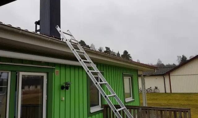 En aluminiumstege lutar mot taket på ett grönt hus, redo för arbete vid skorstenen. En krok till stege är synlig.