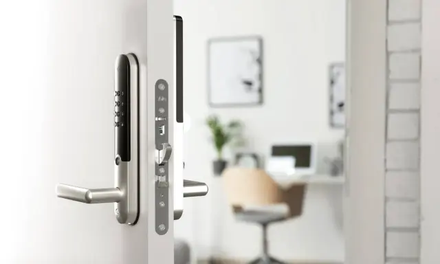 Ett modernt, silverfärgat assa elektroniskt lås med kodpanel och handtag på en vit dörr. I bakgrunden syns ett kontor.