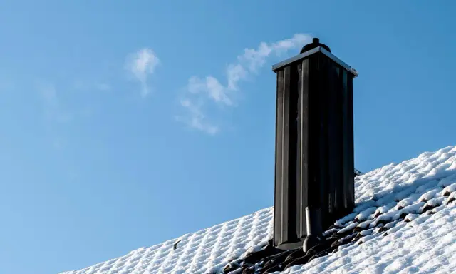 En svart skorsten av plåt ryker på ett snötäckt tak mot en klarblå himmel.