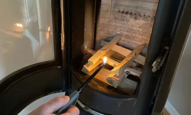 En hand tänder eld i kaminen med en lång tändare. Snart sprider sig värmen i rummet.