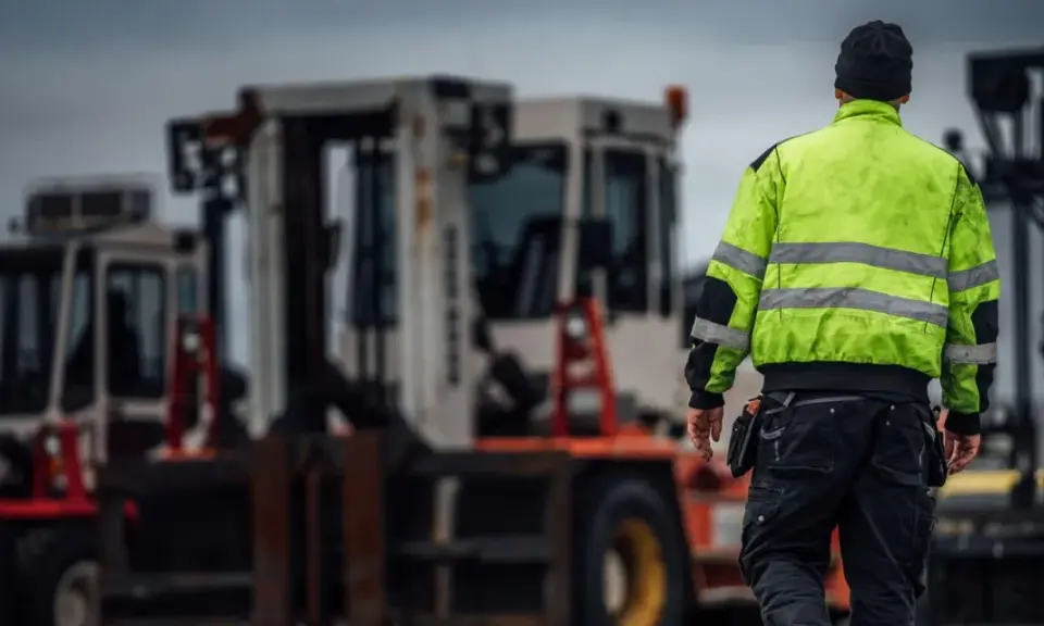 Arbetare i varseljacka inspekterar en truckladdning med flera gaffeltruckar i bakgrunden.