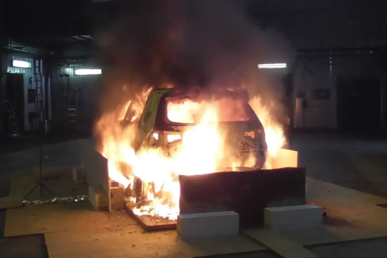 En elbil i ett garage står i full brand. Lågorna slår upp ur bilen och tjock rök fyller rummet. Detta illustrerar brandrisk vid laddning av elbil.