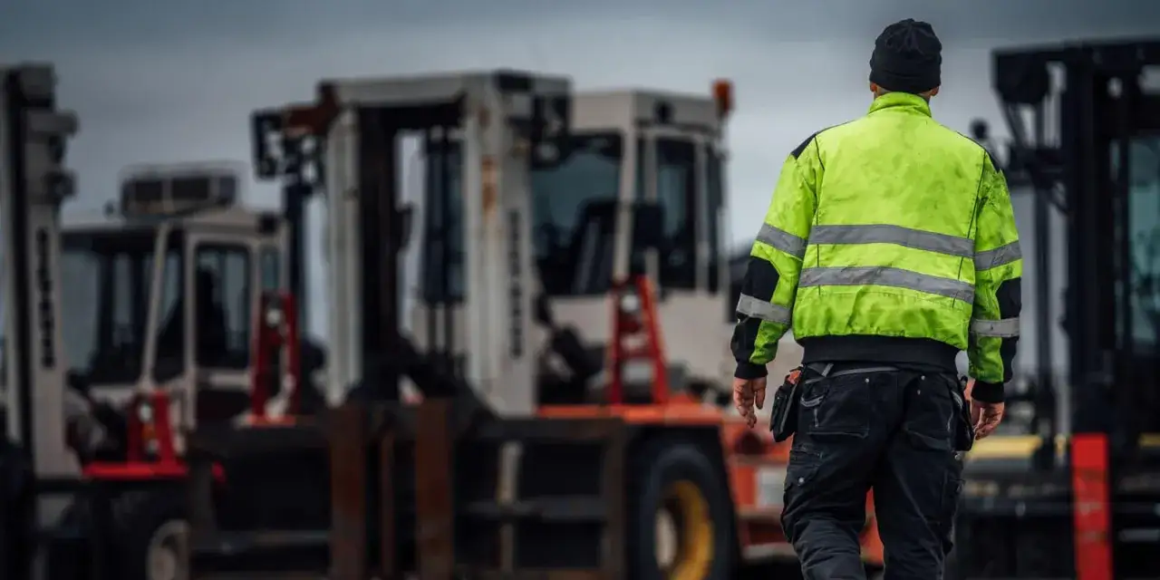 Arbetare i varseljacka inspekterar en truckladdning. Flera truckar st&aring;r uppradade i bakgrunden under en gr&aring; himmel.