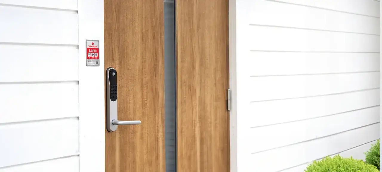 En modern ytterd&ouml;rr med ett Verisure smart l&aring;smodul installation. D&ouml;rren &auml;r i tr&auml; och husfasaden &auml;r vit.