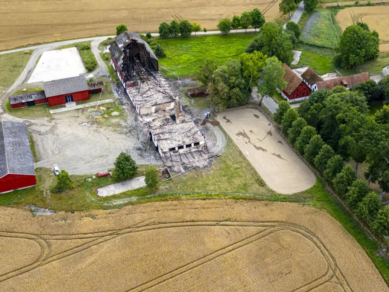 &Ouml;verblicksbild av en g&aring;rd med en delvis f&ouml;rst&ouml;rd stallbyggnad. Omgiven av &aring;kermark och gr&ouml;nska, en ridbana syns ocks&aring;. lbk lantbruk.