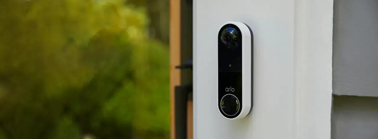 Arlo videod&ouml;rrklocka monterad p&aring; en vit v&auml;gg. Gr&ouml;nskande tr&auml;d syns i bakgrunden.