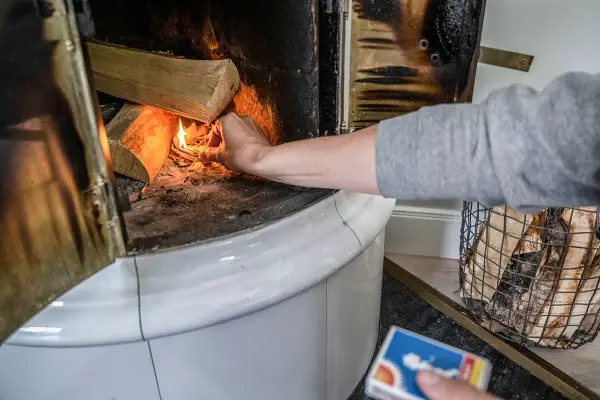 En hand tänder eld i en kakelugn med vedträn som väntar på att värmas.